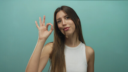 Young hispanic woman with long brown hair makes ok sign in a teal studio set and smiles broadly  approval confidence. © Krakenimages.com
