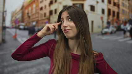 Woman wearing burgundy top points finger to head on busy city street amid blurred traffic; thinking.