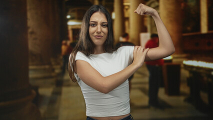 Woman flexes bicep and smiles, raising elbow in a casual white tshirt inside a church building with candle stands and stone pillars; youth empowerment.