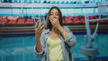 Woman holding smartphone, hand to mouth and fist pump at pool building while smiling broadly; joy celebration.