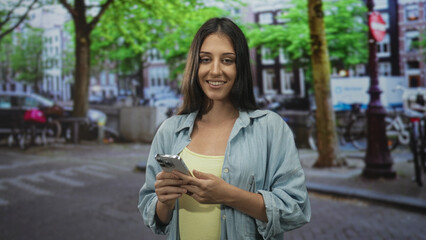 Woman holding smartphone and typing with both hands on a city street while smiling at the screen; digital connection contentment.