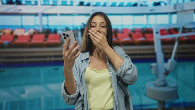 Woman holding smartphone, hand to mouth and fist pump at pool building while smiling broadly; joy celebration.