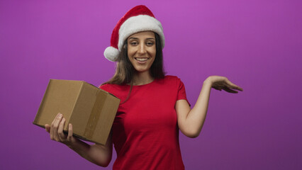 Woman holding cardboard box with palm up gesture in purple studio wearing santa hat and red shirt; holiday cheer.