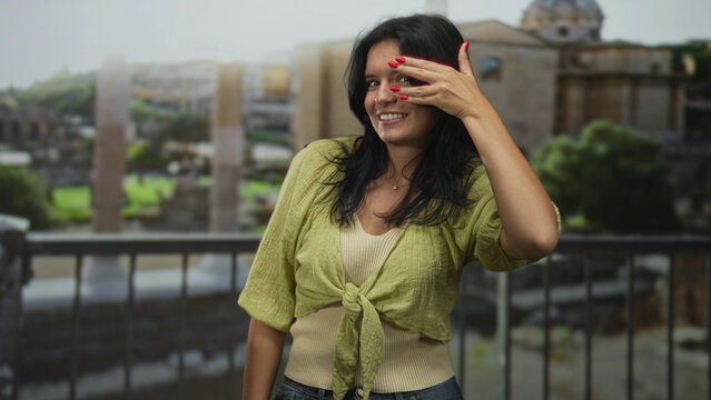Young hispanic woman smiling with hand covering eyes on historic stone balcony of ancient building; shyness.