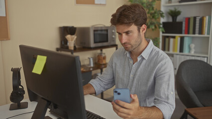 Man holding smartphone and typing on keyboard at desk using desktop computer in building; concentration productivity remote work.