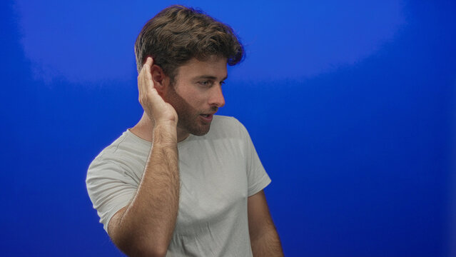 Young hispanic man cupping hand to ear in studio with vivid blue backdrop and white tee; curiosity focus. - Powered by Adobe