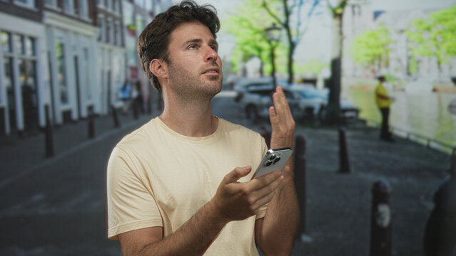 Man holding smartphone with one hand and left hand raised in questioning gesture on a city street; frustration.