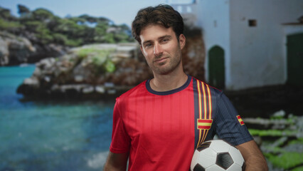 Man holding soccer ball at a seaside building wearing a red team jersey and smiling by the coast; pride.