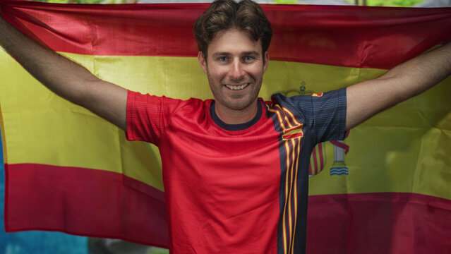 Man smiling with arms raised holding spanish flag and red soccer jersey at resort pool; pride celebration.