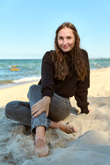 Young woman sitting barefoot on Sandy Beach near sea under clear sky