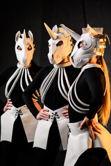 Three performers in futuristic white horse masks stand confidently. Their black outfits highlight the sculpted details of the masks, creating a stylish and conceptual stage atmosphere