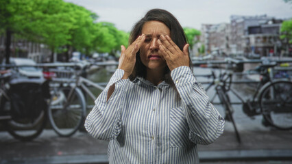 Woman pressing hands to temples and rubbing eyes on a city street by bicycles and canal; fatigue...