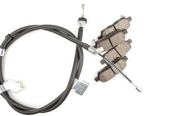 A close-up of new front brake pads with friction material and a metal base. The handbrake cables are encased in a metal, rubberized sheath. White background.