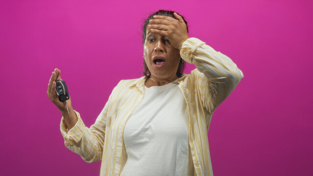 Middleaged hispanic woman holding car key fob with hand on forehead and open mouth in pink studio; panic realization.