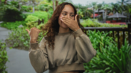 Woman with hand to mouth covering laugh while pointing thumb to self on street walkway lined with green plants and wooden railing; surprise.