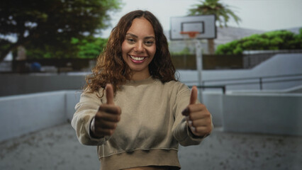 Woman showing two thumbs up while smiling on an outdoor basketball court with hoop visible; joy encouragement.
