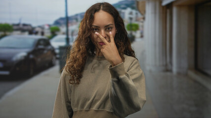 Young african american woman points finger at camera on city street, hand raised to face with visible fingernails and direct gaze; confidence assertion challenge.