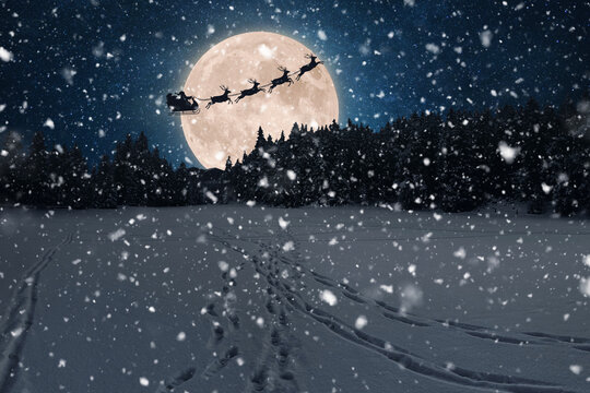 Santa's sleigh and reindeer fly silhouetted across a giant full moon in a magical, snowy night sky. Fresh snow falls over pristine winter landscape with footprints. Christmas and winter holiday themes