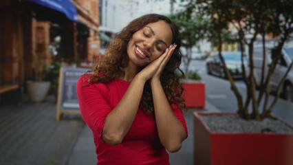 Fototapeta premium Young african american woman with hands together and cheek resting in sleeping gesture on busy city street; restful serenity.