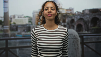 Fototapeta premium Woman showing face and smiling while standing near ancient roman ruins at a building railing outdoors wearing striped top; serenity travel memory.