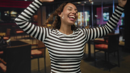 Woman dancing with arms raised and smiling in a striped shirt inside a restaurant building, head tilted back and eyes closed; joy.