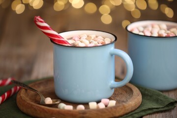 Tasty cocoa with marshmallows and candy cane on wooden table against blurred Christmas lights, closeup