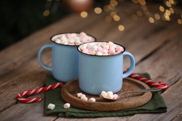 Tasty cocoa with marshmallows and candy canes on wooden table against blurred Christmas lights, closeup
