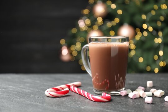 Tasty hot cocoa, marshmallows and candy canes on black table against blurred Christmas lights