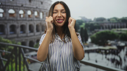 Fototapeta premium Hispanic woman with clenched fists near face smiling by the coliseum building on a tourist balcony, outdoor scene; joy travel memory.