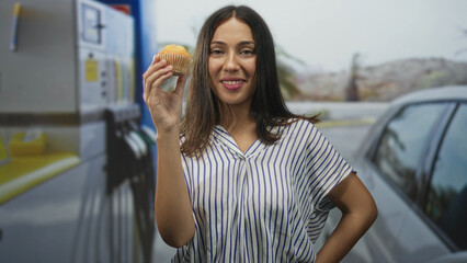 Woman holding muffin with bare hand at street petrol station, smiling toward camera; cheerful snack moment.