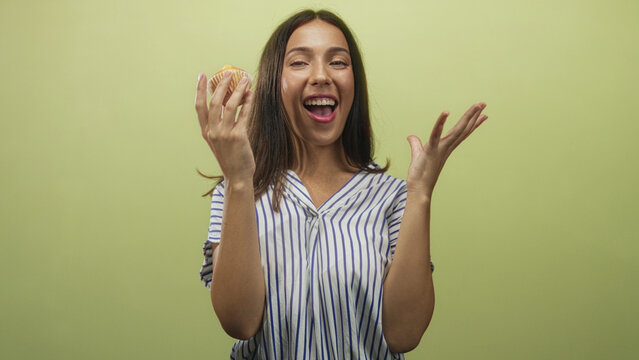 Hispanic young brunette woman holding cupcake with raised hands and smiling in green studio wearing striped shirt; joy celebration.