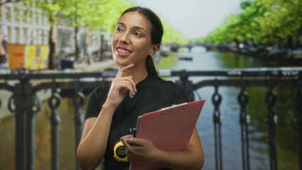 Hispanic woman referee with whistle and stopwatch holding clipboard, finger to chin for thinking on a street in amsterdam; thoughtful planning.