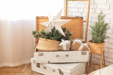Vintage white suitcases with Christmas gifts, holiday paper star decor and a basket with fir branches in a cozy home interior design. Festive winter decoration setup in a warm living room at home