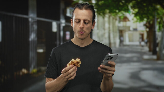 Man holding waffle and smartphone on street, looking at phone while eating and scrolling; digital distraction contemplation.