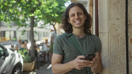 Man texting on a smartphone while leaning against a building wall on a busy city street, shoulder bag strap visible; contemplation.