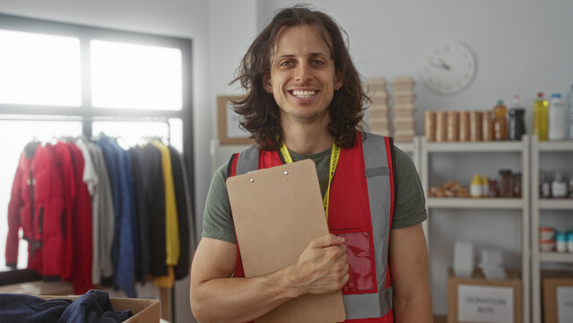 Man holding clipboard and smiling while wearing a volunteer vest in a donation building with coats and boxes; helpful community service spirit.