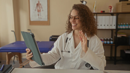 Woman doctor with stethoscope smiling and waving hand while holding tablet in building clinic exam room; compassion telemedicine.