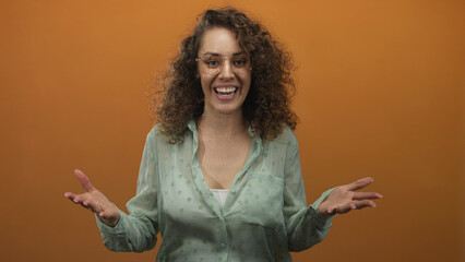 Woman wearing green polka dot blouse and glasses smiling with raised hand gesture in studio with orange backdrop; excitement playfulness warmth.