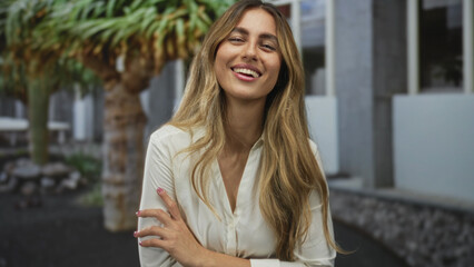 Young blonde woman bare arms smiling with hands on hips in stone courtyard surrounded by potted plants; confidence joy warmth.