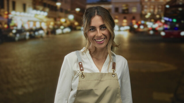 Woman in apron smiles showing face in city street with blurred lights behind at night; friendliness greeting.