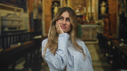 Young slender blonde woman holds bare hand on cheek in historic cathedral; toothache pain worry anxiety.