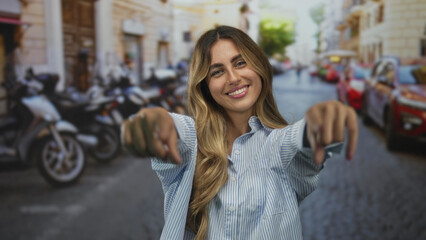 Young blonde woman with flowing hair hand pointing to camera while smiling brightly in sunlit european city street; joy confidence.