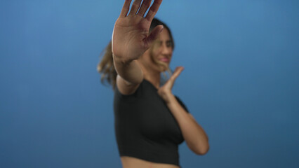 Blonde woman holds up open hand in stop gesture with facial grimace in photo studio against blue...