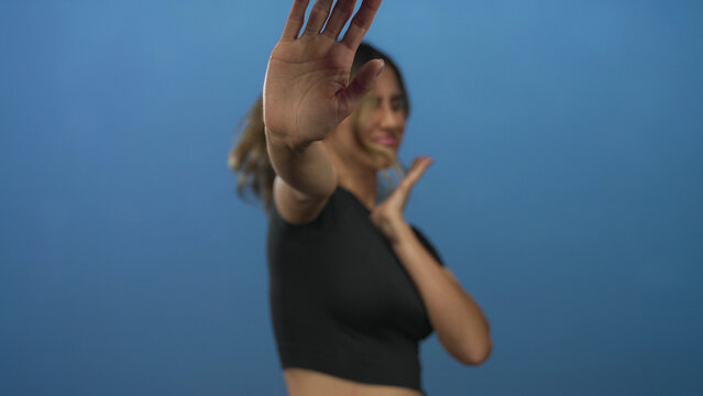 Blonde woman holds up open hand in stop gesture with facial grimace in photo studio against blue backdrop; rejection discomfort.