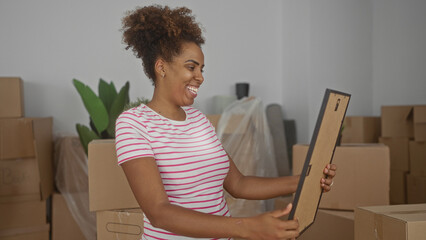 Woman holding a picture frame, smiling with hands visible among stacked moving boxes inside a house; moving day joy.