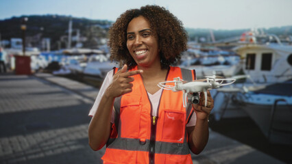 Woman pointing finger to drone at port marina beside boats wearing orange safety vest holding quadcopter; confidence innovation.