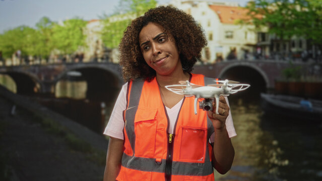 Young woman technician holding drone in hand on street in amsterdam canal, wearing orange safety vest while examining quadcopter; frustration. - Powered by Adobe