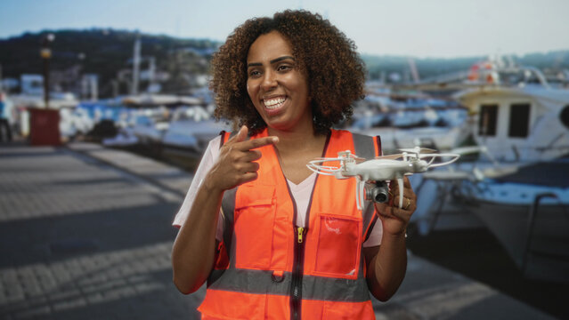 Woman pointing finger to drone at port marina beside boats wearing orange safety vest holding quadcopter; confidence innovation.
