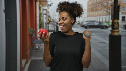 Woman smiling holding apple in left hand and dumbbell in right hand on city street wearing black shirt; health and happiness.
