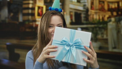 Woman wearing a party hat holds a blue gift box inside a candlelit building while smiling; happiness.
